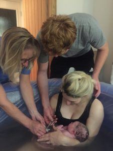 Charina is holding her newborn baby in a birth pool as her husband cuts the cord, assisted by Sue the midwife