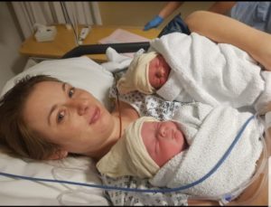 Carla is holding her newborn twins wrapped in blankets wearing hats. She is lying on a hospital bed smiling.